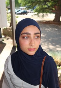 Marineblauer Hijab, glatte Textur, kombiniert mit einem weißen Kleidungsstück. Hat klare Haut und grüne Augen, mit einem braunen Riemen, der auf der Schulter sichtbar ist.