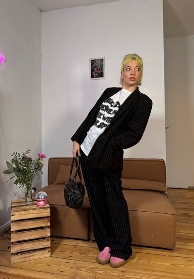 Traje negro a rayas con una camiseta gráfica blanca, zapatos de plataforma rosas y un bolso negro. El fondo incluye un sofá marrón y un jarrón con flores.