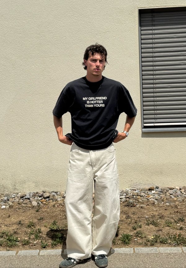 Jeune homme portant un T-shirt noir avec du texte blanc "MY GIRLFRIEND IS HOTTER THAN YOURS", debout contre un mur beige près d'une fenêtre.