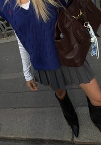 Suéter azul de malha, camisa branca de manga longa, saia plissada cinza, botas pretas até ao joelho e uma mala de mão em pele castanha com detalhes em metal.