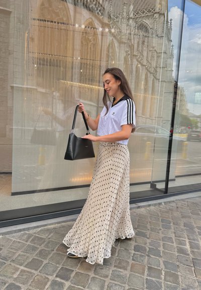 Camisa blanca de manga corta con rayas negras, combinada con una falda beige de lunares fluida; sosteniendo un bolso negro, de pie sobre adoquines.