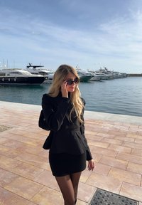 Blazer preto ajustado sobre uma minissaia preta canelada, combinado com collants pretos transparentes e óculos de sol oversized, em um cenário de marina.