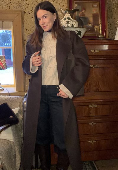 Mujer de cabello oscuro que lleva un abrigo largo y oscuro, suéter de punto grueso color crema y pantalones oscuros en una habitación de estilo vintage con muebles de madera.