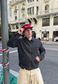 Fekete-fehér csíkos dzseki cipzáras záródással és elülső zsebbel, piros sapkával, melyen fehér felirat található, városi környezetben.