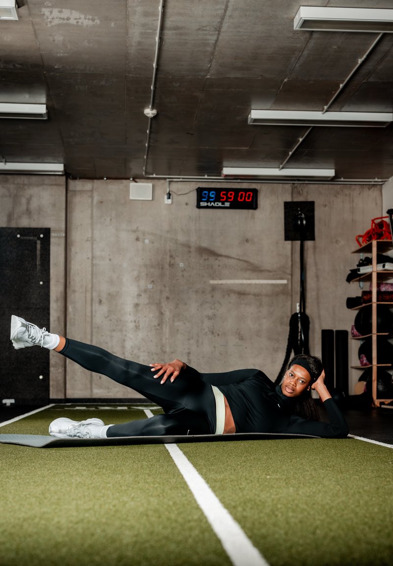 Femme en tenue de sport noire s'exerçant sur le côté avec la jambe levée sur un tapis en gazon vert dans une salle de sport avec un minuteur au mur.