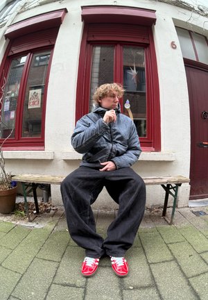 Una persona se sienta en un banco de madera con una chaqueta acolchada oscura, pantalones negros holgados y zapatillas rojas con cordones azul claro, frente a un edificio.