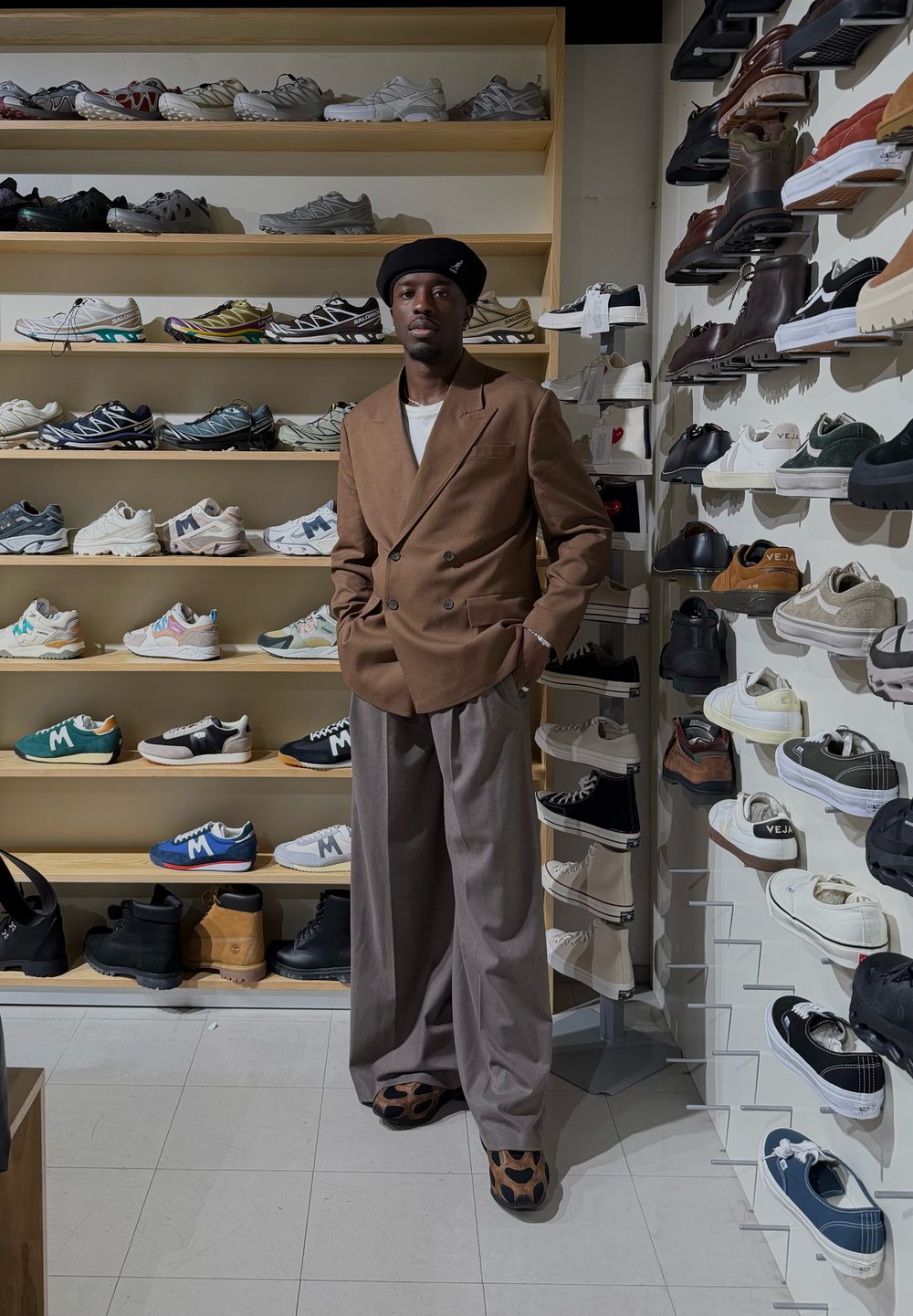 Brauner zweireihiger Blazer, weißes Hemd und hellgraue Hose. Steht auf dem Boden eines Schuhgeschäfts mit Regalen voller verschiedener Sportschuhe im Hintergrund.