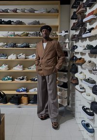 Brauner zweireihiger Blazer, weißes Hemd und hellgraue Hose. Steht auf dem Boden eines Schuhgeschäfts mit Regalen voller verschiedener Sportschuhe im Hintergrund.