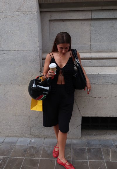 Top corto negro con lazo en la parte delantera, pantalones cortos negros, zapatos planos de malla rojos. Sosteniendo una taza de café, un casco negro y una bolsa de compras amarilla.