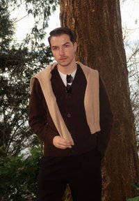 Νεαρός άνδρας που φοράει σκούρο καφέ πουλόβερ με μπεζ πουλόβερ ριγμένο στους ώμους, στέκεται δίπλα σε ένα δέντρο σε ένα δασώδες εξωτερικό περιβάλλον.