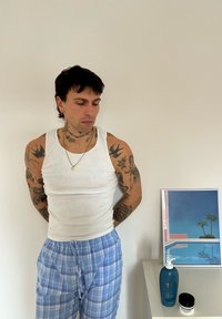 Witte geribbelde tanktop, blauwe geruite pyjamabroek, tatoeages op armen en hals. Naast een ingelijste print en huidverzorgingsproducten op een witte tafel.