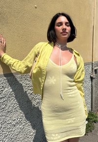 Jasnożółta dzianinowa sukienka w kwiatowe wzory, zestawiona z przezroczystym żółtym kardiganem. Gładka tekstura, dopasowany krój i minimalne akcenty metalowe.