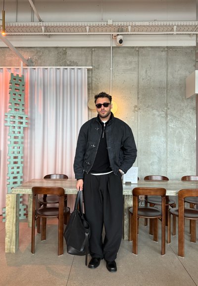 Hombre con chaqueta negra y pantalones anchos sosteniendo una bolsa negra se encuentra en un comedor moderno con sillas de madera y una pared de cemento de fondo.