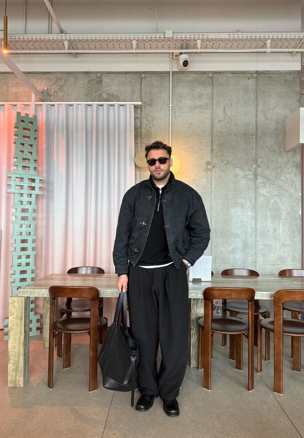 Mann in schwarzer Jacke und weiten Hosen, der eine schwarze Tasche hält, steht in einem modernen Essbereich mit Holzstühlen und Betonwand im Hintergrund.