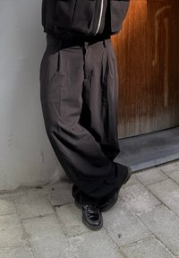 Φαρδιές μαύρες παντελόνα με πιέτες, φτιαγμένες από απαλό ύφασμα, συνδυασμένες με γυαλιστερές μαύρες παπούτσια, τοποθετημένες σε μία υφή φόντου.