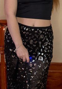 Jupe noire à sequins avec une surcouche transparente, ornée de sequins circulaires et une taille ajustée. Une main tient une lanière noire et un petit objet bleu.