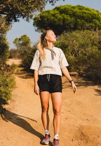 Lys beige t-skjorte med halsduk, svarte shorts og grå-lilla joggesko. Utendørsmiljø med sti og grøntområder synlig.