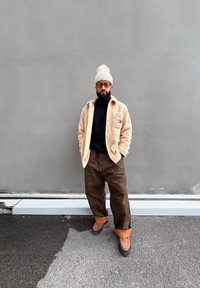 Μπεζ φλιτζάνι μπουφάν πάνω από μια μαύρη ζακέτα, καφέ καρό παντελόνι, μπεζ σκούφος, κίτρινα γυαλιά ηλίου και καφέ παπούτσια. Στηρίζεται σε έναν γκρίζο τοίχο.