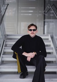 Pull noir à col roulé, pantalons larges noirs avec des rayures jaunes, chaussures noires, lunettes de soleil, assis sur des escaliers gris avec une rambarde en verre.