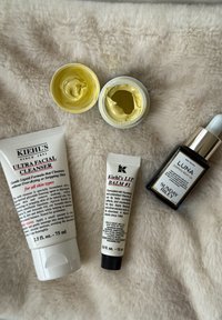 Kiehl's Ultra Facial Cleanser in einer weißen Tube, Kiehl's Lip Balm #1 in einer schwarzbedoppten Tube und zwei gelbe Cremetöpfe, alle auf einem weichen Hintergrund.
