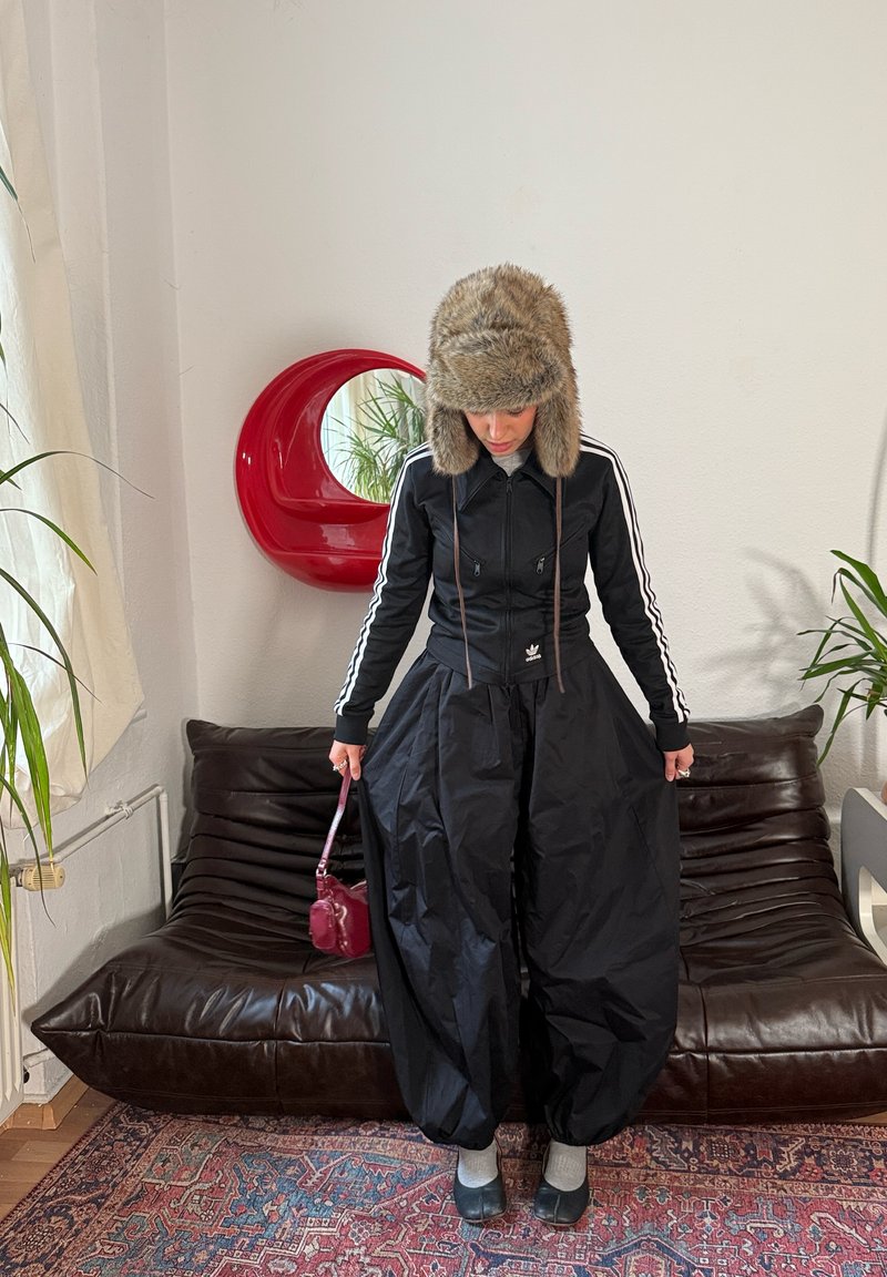 Coupe-vent noir avec bandes blanches, pantalons noirs surdimensionnés, chaussettes grises et chapeau en fourrure ; tenant un petit sac bordeaux. Miroir circulaire rouge en arrière-plan.