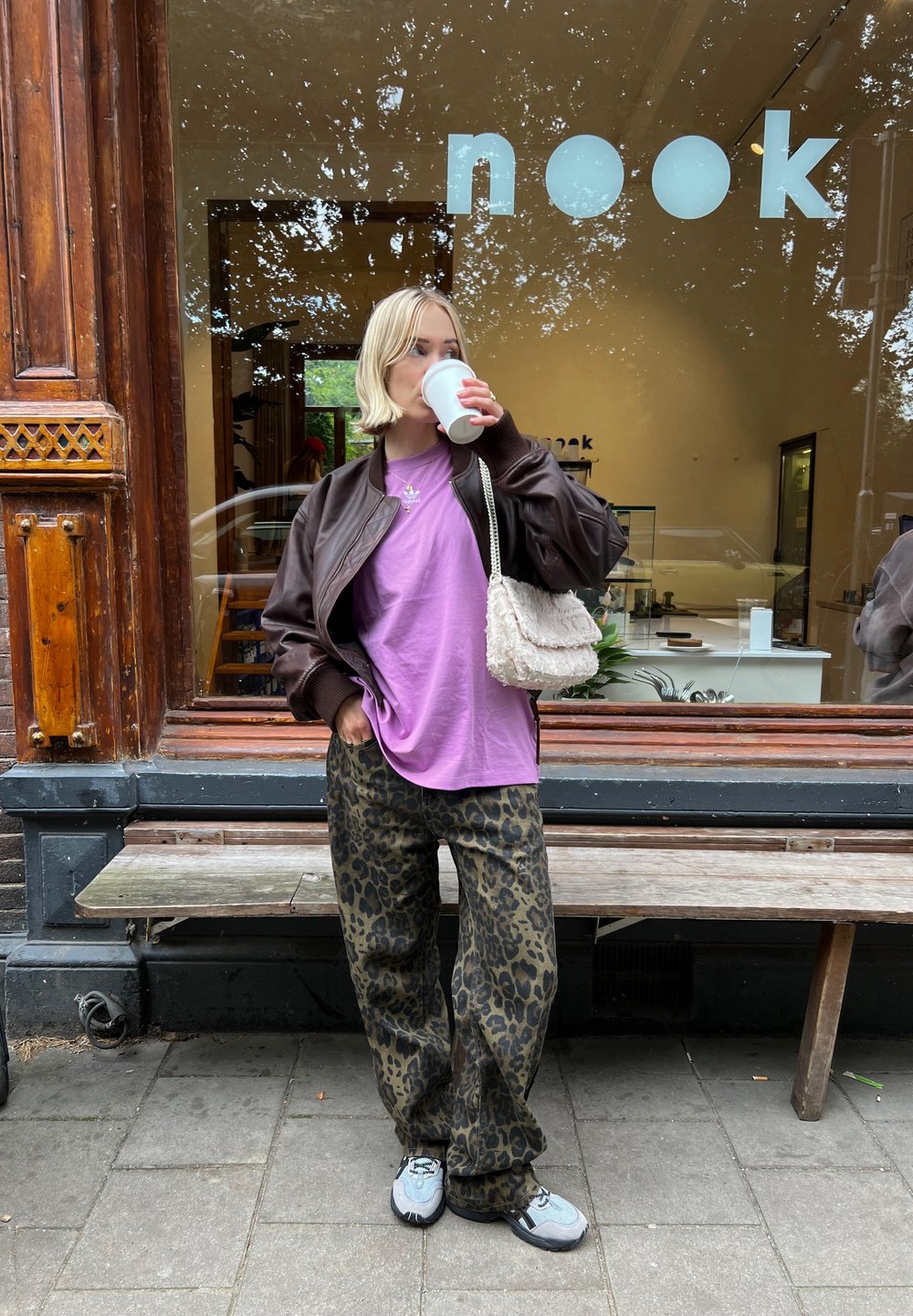 Jachetă bomber maro peste un tricou mov, asortată cu pantaloni verzi cu imprimeu leopard și adidași gri. Ținând o cană de cafea, stând în aer liber.
