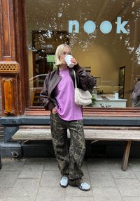 Jachetă bomber maro peste un tricou mov, asortată cu pantaloni verzi cu imprimeu leopard și adidași gri. Ținând o cană de cafea, stând în aer liber.