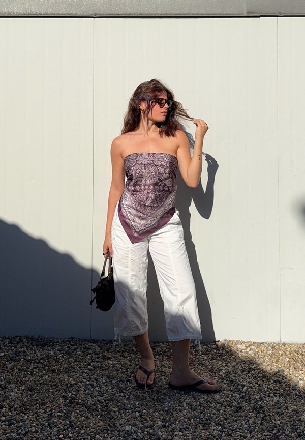 Femme en pantalon court blanc et haut tube violet à motifs se tient sur du gravier, tenant un sac à main noir et touchant ses cheveux contre un mur blanc.