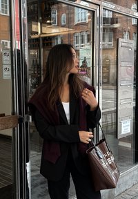 Μαύρο σακάκι επάνω από ένα λευκό μπλουζάκι με μαρόν πουλόβερ ριγμένο στους ώμους. Κρατάει μια καφέ τσάντα tote με λευκό κείμενο.