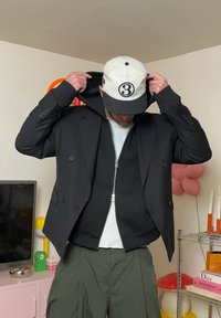 Svart dobbeltspent jakke over en svart zip-up hettegenser, båret med en hvit ribbet skjorte og løse olivengrønne bukser. Hvit og svart caps.