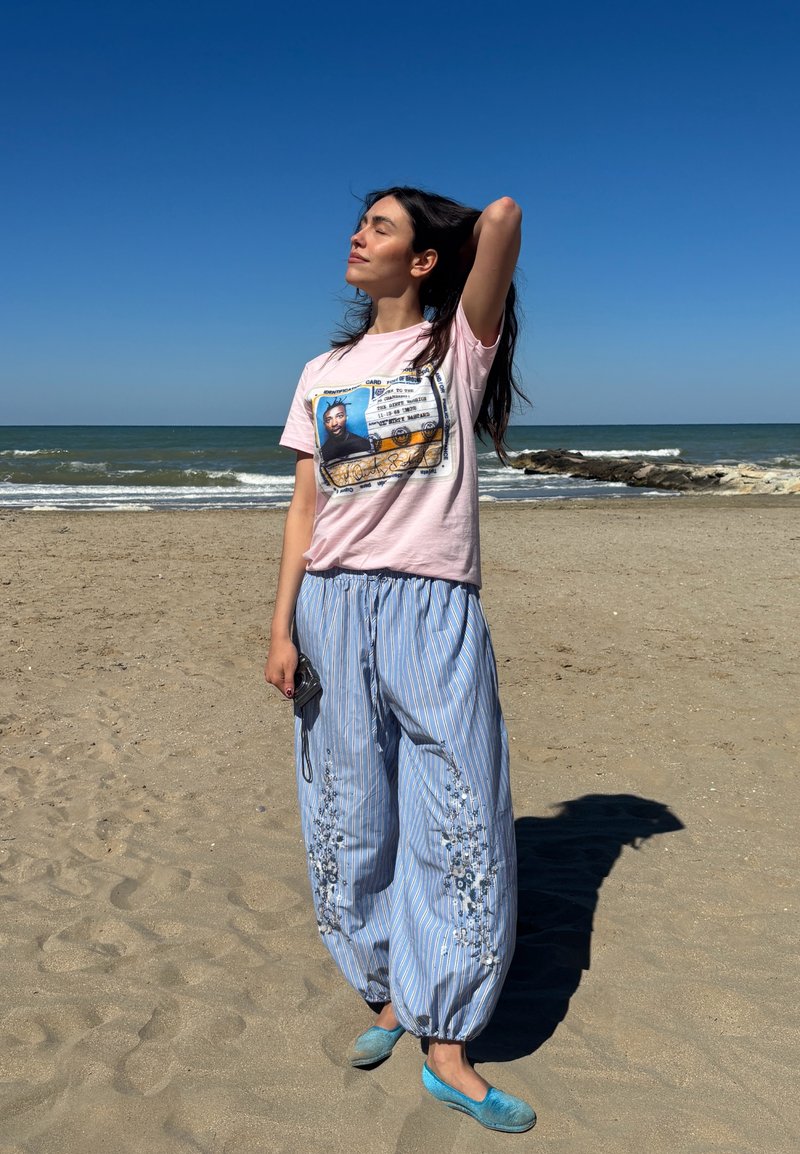 Femme se tient sur une plage de sable près de l'océan, vêtue d'un pantalon bleu rayé, d'un t-shirt rose à motif graphique et des chaussures bleues, les yeux fermés et la main dans les cheveux.