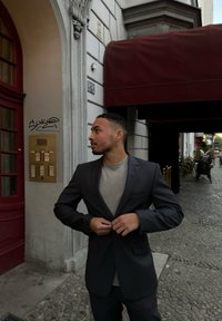Osoba ubrana w ciemnoszary żakiet na jasnoszarym koszuli stoi obok czerwonych drzwi z mosiężnym domofonem i kamiennym chodnikiem.
