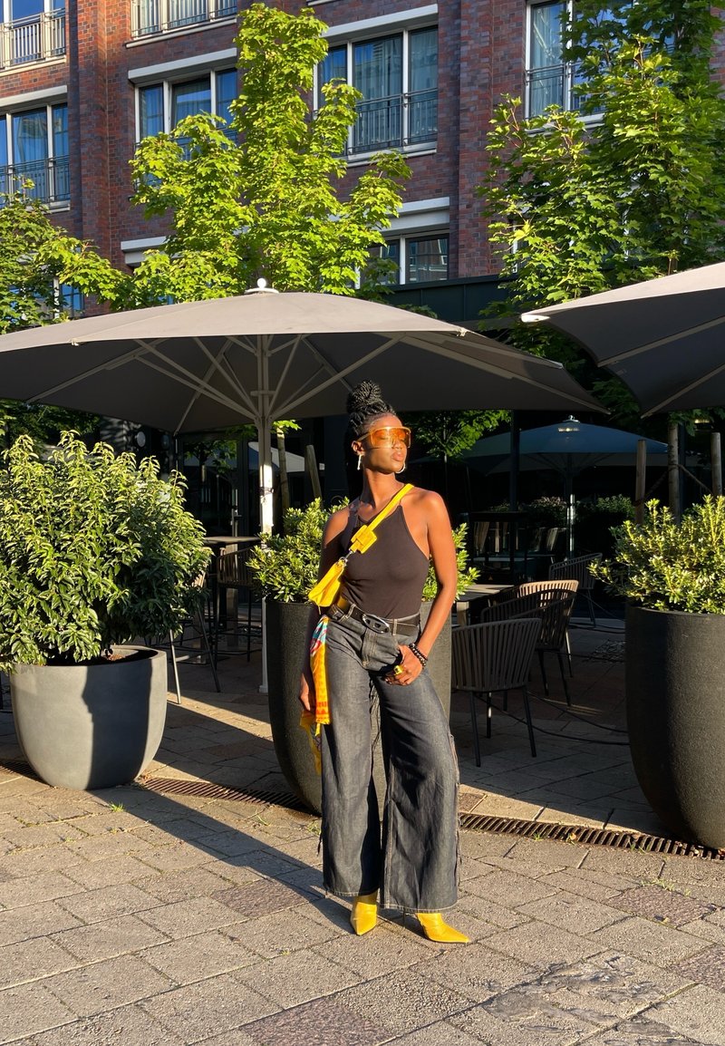 Schwarzes Tanktop, hoch-taillierte dunkle Jeans mit weitem Bein, gelbe Absätze, gelbe Taschenriemen, Sonnenbrille, Grünpflanzen und Sonnenschirme im Hintergrund.