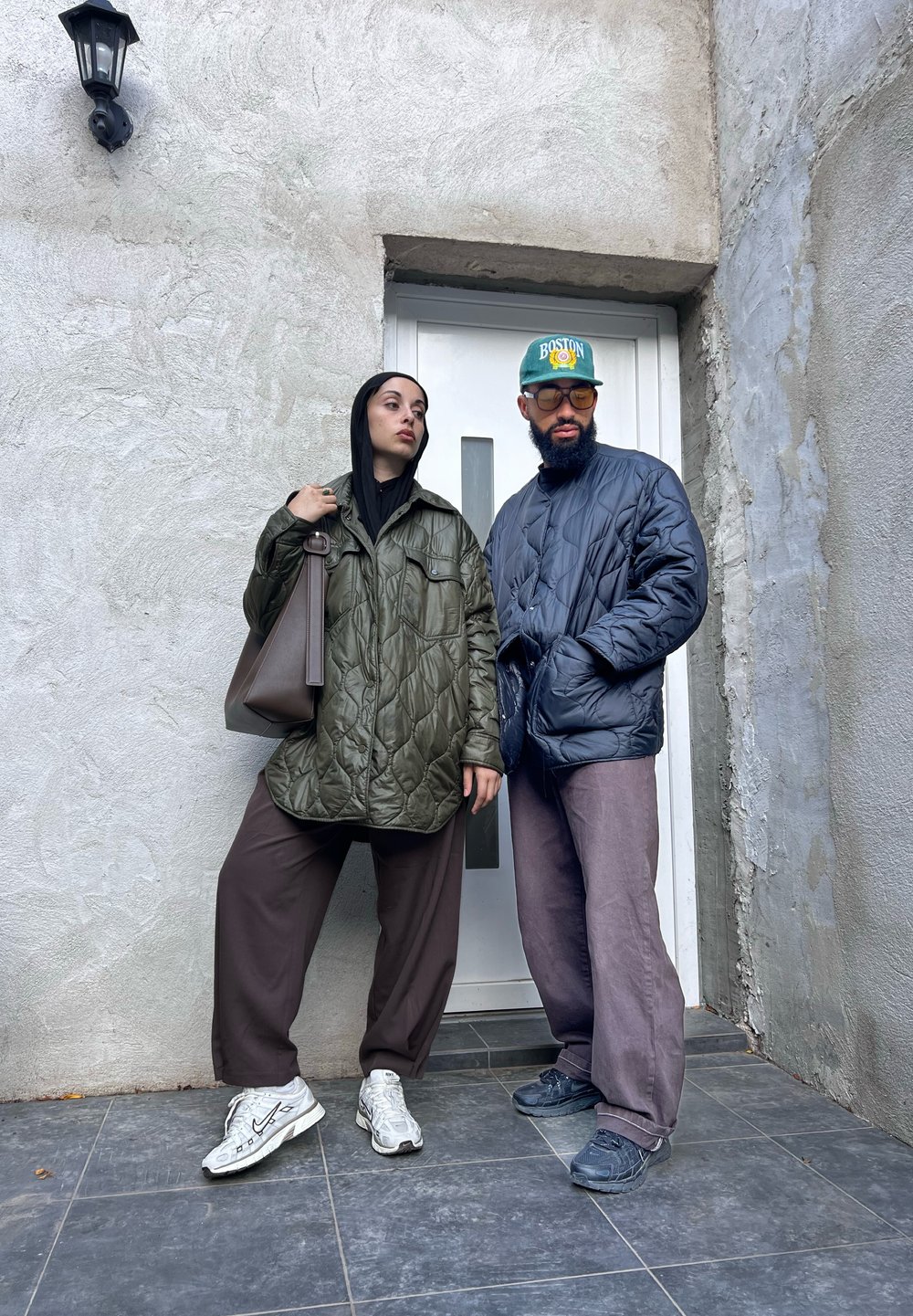 Κουβερτίνα ελαιόλαδου πράσινη ζακέτα, σκούρα κουβερτίνα ζακέτα, καφέ παντελόνια και αθλητικά παπούτσια. Υπαίθριο περιβάλλον με υφή γκρι τοίχου και πόρτας.