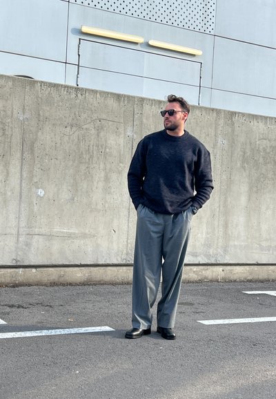 Pantalones de pierna ancha grises, suéter oscuro y zapatos negros. De pie contra una pared de concreto, con los brazos relajados a los lados, usando gafas de sol oscuras.