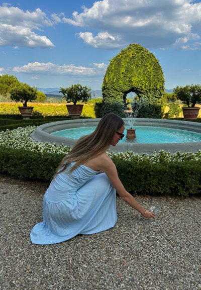 Vestido sin mangas de color azul claro con un acabado texturizado. Persona arrodillada junto a un camino de grava, extendiendo la mano hacia un vaso cerca de una fuente circular.