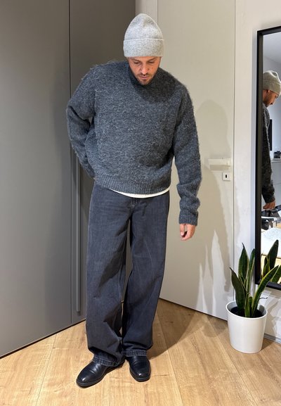 Hombre con gorro de punto gris, suéter esponjoso gris oscuro, pantalones vaqueros negros holgados y zapatos negros, de pie en el interior cerca de un espejo y una planta en maceta.
