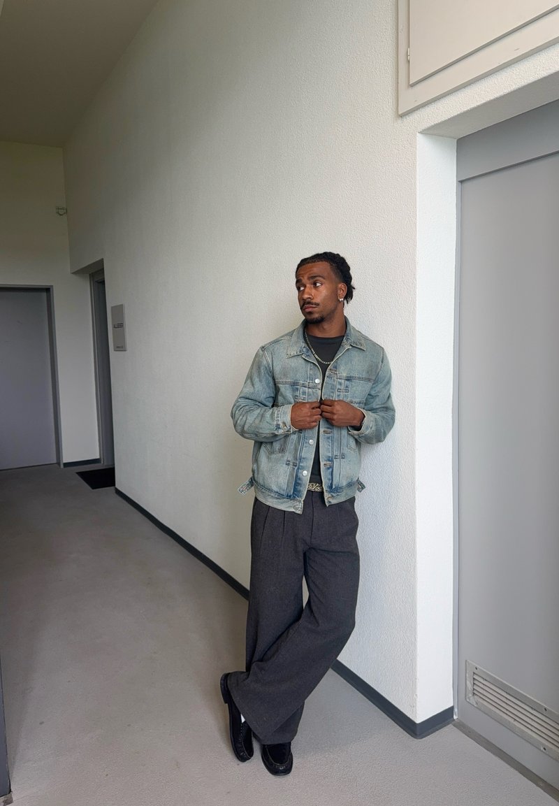 Homme en veste en jean et pantalons larges foncés, appuyé contre un mur blanc dans un couloir avec des portes grises, regardant vers sa gauche.
