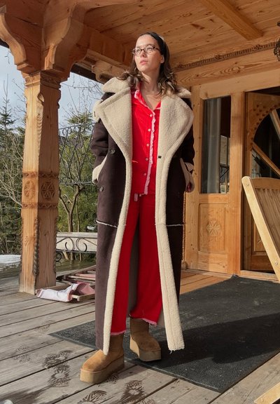 Abrigo de lana marrón con cuello de piel sintética crema, camisa de cuadros roja, y botas de tobillo color beige. Fondo de porche de cabaña de madera con plantas.