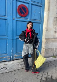 Γυναίκα με μαύρο τοπ με πουά, κόκκινο κασκόλ, δαντελωτή φούστα πάνω από μαύρο παντελόνι, κρατώντας κίτρινη τσάντα, στέκεται μπροστά σε μια μεγάλη μπλε πόρτα με πινακίδα απαγόρευσης στάθμευσης.