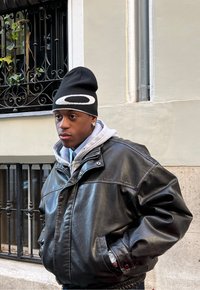 Veste bomber en cuir noir avec un grand col sur un hoodie gris. Bonnet noir avec logo blanc. Brillance subtile sur la texture de la veste.