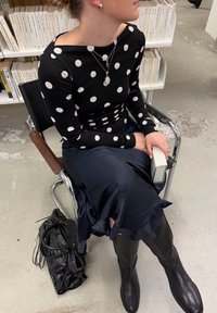 Γυναίκα με μαύρο τοπ με πουά και μπλε φούστα κάθεται σε καρέκλα κρατώντας ένα βιβλίο, με μια μαύρη τσάντα στο πάτωμα κοντά στα πόδια της.