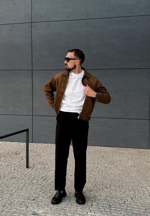 Chaqueta de ante marrón sobre una camiseta blanca, pantalones negros y mocasines negros, de pie contra una pared gris con superficie texturizada.