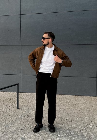 Chaqueta de ante marrón sobre una camiseta blanca, pantalones negros y mocasines negros, de pie contra una pared gris con superficie texturizada.