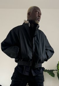 Czarna kurtka bomberka z pluszowym kołnierzem, elastycznym pasem i błyszczącym wyko�ńczeniem; zestawiona z ciemną koszulą i fakturowanymi czarnymi spodniami.