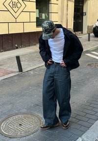 Calças de ganga de perna larga com lavagem escura, t-shirt branca, casaco preto e um boné camuflado. Sapatos castanhos com detalhes brancos apresentam uma sola robusta. Fundo de passeio.