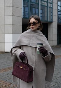 Manteau en laine beige avec un large col et une fermeture à boutons, écharpe grise, sac à main en daim bordeaux, gants marron et lunettes de soleil marron surdimensionnées.