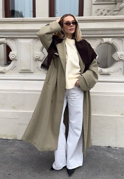 Mujer con abrigo beige extragrande, suéter crema, pantalones amplios blancos, zapatos negros puntiagudos y gafas de sol, de pie frente a una pared de piedra ornamentada.