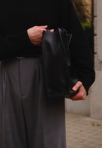 Černá kožená taška s měkkou texturou, obdélníkového tvaru a zapínáním na zip, držená v ruce vedle šedých kalhot.