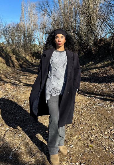 Abrigo negro largo sobre un suéter texturizado gris claro, combinado con pantalones grises y botas marrones; ambientado en un entorno natural al aire libre.